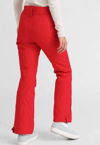 Pantalon imperméable rouge avec une taille élastique, des jambes droites et des aérations zippées aux chevilles ; assorti à des baskets gris clair.
