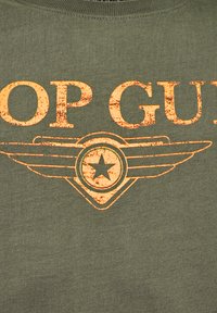 Olivgrön bomull T-shirt med en sliten orange "TOP GUN" text och en stjärna med vinge-design i mitten.