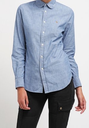 Kobieta ubrana w jasno-niebieską koszulę na guziki z długim rękawem oraz czarne spodnie z boczną kieszenią, stojąca na jednolitym tle.