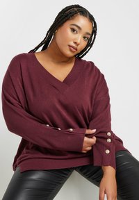 Burgunderfarbener Oversized-Pullover mit V-Ausschnitt, gerippter Textur und dekorativen silbernen Knöpfen an den Ärmeln, kombiniert mit schwarzen, figurbetonten Hosen.