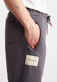 Graue Sweatpants mit Taschen; Merkmale ein Stoffetikett mit der Aufschrift "STUDIO EDITION" in Creme, elastischer Bund mit Kordelzug, glatte Textur.