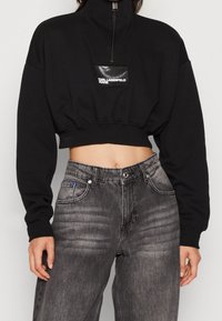 Beskarvad svart sweatshirt med hög krage och dragkedja, med en blank svart logotyp. Matchas med högmidjade grå jeans.