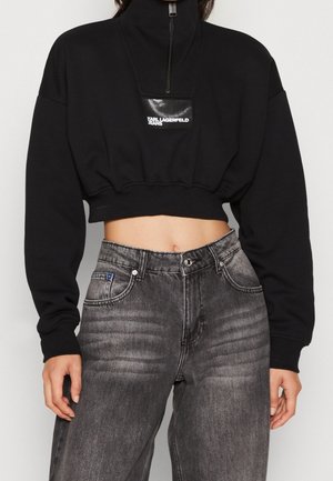 Sudadera negra recortada con cuello alto y cierre de cremallera, con un parche de logo negro brillante. Combinada con jeans grises de talle alto.