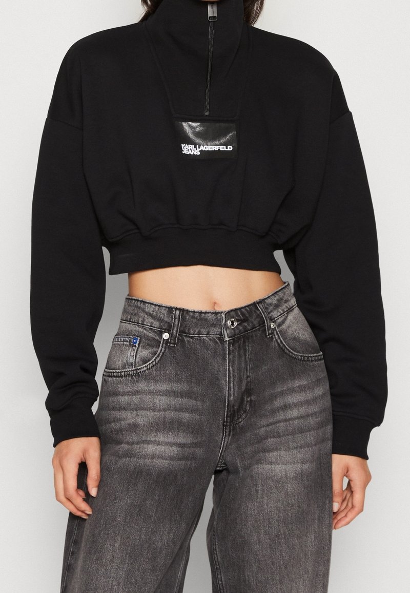 Beskarvad svart sweatshirt med hög krage och dragkedja, med en blank svart logotyp. Matchas med högmidjade grå jeans.