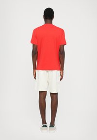 Red short-sleeve t-shirt with a crew neck, worn with white shorts and green-accented shoes, viewed from the back.
