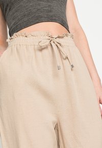 Pantalon en tissu beige léger avec taille élastique, bordure à volants et fermeture à cordon. Comprend deux poches latérales.