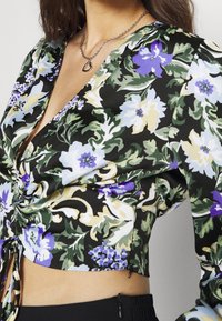 Top corto floreale nero con grandi fiori blu, viola e crema accompagnati da fogliame verde; tessuto morbido, scollatura a V e dettaglio arricciato sui lati con lacci.