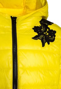 Gul puffjacka med dragkedja fram, med en svart broderad blommönster på axeln och en glansig, slät textur.