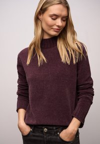 Pullover in tiefem Violett, strukturierter Stoff mit hohem Kragen und gerippten Bündchen. Getragen mit den Händen in den Taschen, schwarze Jeans sichtbar.