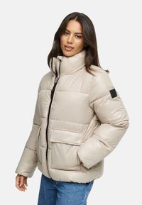 Beige gewatteerde jas met hoge kraag, ritssluiting en twee voorzakken. Heeft een zwart logopatch op de mouw. Effen, glanzende textuur.