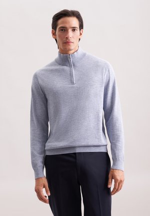 Mann in einem hellgrauen Reißverschluss-Pullover mit hohem Kragen und dunklen Hosen, der vor einem schlichten hellen Hintergrund steht.