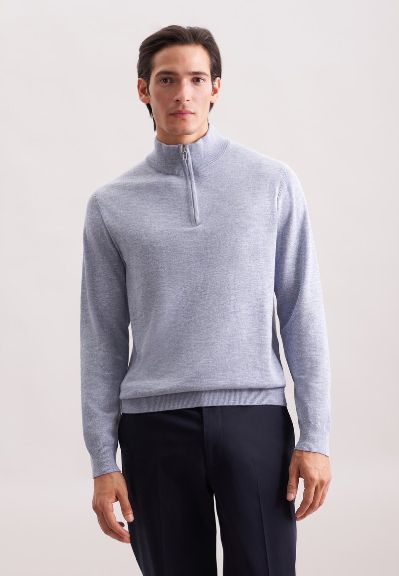 Homme portant un pull zippé gris clair avec un col haut et un pantalon sombre, se tenant devant un fond clair.