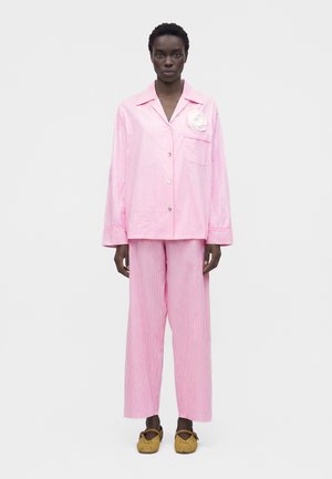 Model trägt lockeren, rosa-weißen Pyjama mit vertikalen Streifen, weißer Blumenbrosche und hellbraunen geflochtenen Mary Jane Schuhen vor weißem Hintergrund.