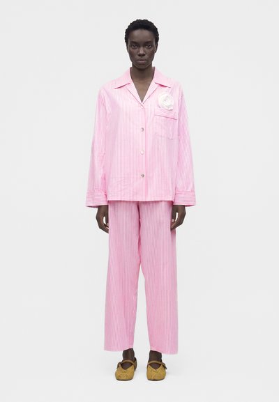 Mannequin portant un ensemble pyjama ample à rayures verticales rose et blanc avec une broche florale blanche et des chaussures Mary Jane tissées couleur fauve sur fond blanc.