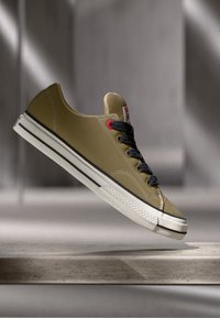 Khaki sneaker with a smooth, synthetic upper, black laces, white sole, and red accents. Features a round toe and classic low-top silhouette.