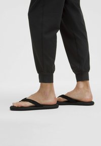 Schwarze Flip-Flops mit einem breiten Riemendesign und glatten Sohlen, getragen zu dunklen, schmal geschnittenen Hosen, die einen minimalistischen Stil zur Schau stellen.