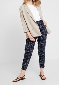 Beige dvouřadový blazer, bílá košile, námořnicky modré kalhoty s puntíky a hnědé sandály se strappy pásky, vše v uvolněném střihu a s ohrnutými rukávy.