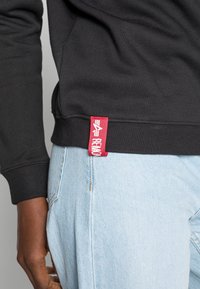 Sweat-shirt noir avec poignets et ourlet côtelés, arborant une étiquette tissée rouge. Associé à un jean bleu clair affichant une coupe décontractée et une texture apparente.