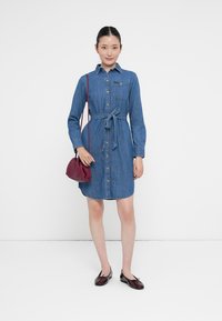 Denim skjortekjole med lange ermer, knappelukking foran, belte i midjen og brystlomme; brukt med en liten burgunder håndveske og flate sko.