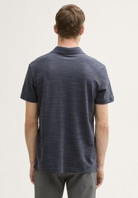 Koszulka polo z krótkim rękawem w ciemnym granacie z teksturowanym, subtelnie paskowanym wzorem. Posiada klasyczny kołnierzyk oraz prosty dół, wykonana z miękkiego materiału.