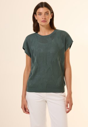 Dunkelgrüner Strickpullover mit strukturiertem, gemustertem Design. Kurze Ärmel und ein Rundhalsausschnitt, kombiniert mit weißen Hosen.