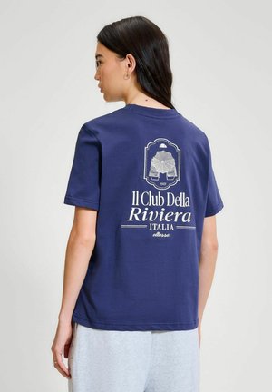 Mujer con cabello oscuro que lleva una camiseta azul marino con el texto blanco "Il Club Della Riviera Italia" y un gráfico en la espalda.