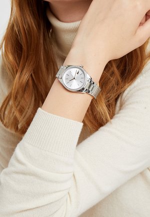 Montre-bracelet en argent avec un cadran blanc rond, des marqueurs minimalistes et une lunette ornée de cristaux ; attachée à un bracelet en métal poli.