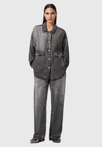 Mujer de pie vistiendo una chaqueta de mezclilla negra desgastada y oversized con botones plateados y pantalones vaqueros de pierna ancha a juego, contra un fondo liso.
