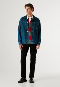 Giacca di jeans blu con chiusura a bottoni, tasca frontale, indossata su una maglietta a righe grigia e rossa. Pantaloni neri e scarpe nere completano l'outfit.