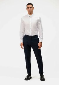 Camicia bianca con bottoni, taglio slim, maniche lunghe; abbinata a pantaloni su misura blu scuro e scarpe nere. Tessuto liscio, design minimalista.