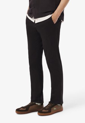 Mann trägt schwarze Hose, schwarze, geknöpfte Weste über weißem Hemd und braune Turnschuhe mit grünem Logo, steht mit den Händen in den Taschen.