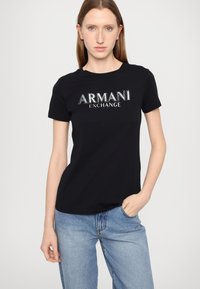 Svart bomullst-skjorte med kort erme som har en sølvfarget "ARMANI EXCHANGE" logo over brystet. Brukes med lyseblå jeans.