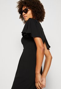 Svart kjole med korte, volangermed ermer, tettsittende bodice og flytende skjørt. Modellen har på seg svarte solbriller, som viser frem strukturert hår.