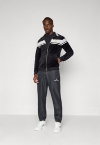 Svart velour treningsjakke med hvite striper og gullaccenter, kombinert med grå bukser med elastisk midje som har et logo, iført hvite sneakers.