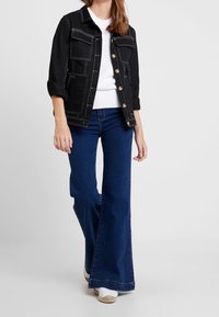 Schwarze Jeansjacke mit weißen Nähten, Knopfverschluss und vier Fronttaschen, kombiniert mit weit geschnittenen dunkelblauen Jeans und weißen Sneakern.