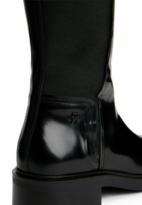 Black polished leather ankle boot with textured fabric upper section, block heel, side zipper, and small embossed brand logo near heel.