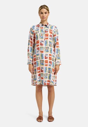 Frau trägt ein knöpfbares, langärmeliges Kleid mit buntem Sardinendosenmuster, steht mit entspannten Armen vor weißem Hintergrund.