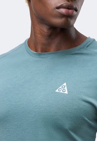 Κοντινό πλάνο ενός ατόμου που φοράει ένα ανοιχτό μπλε T-shirt με ένα μικρό λευκό τρίγωνο λογότυπο "acg" στο αριστερό στήθος.