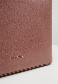 Gros plan d'un sac Ted Baker en cuir texturé marron avec le nom de la marque embossé en or dans le coin inférieur gauche.