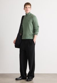 Green zip-up sweater with a small logo, black pleated pants, and black shoulder bag, set against a neutral background. Smooth fabric texture.