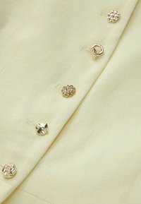 Nahaufnahme eines blassgelben Kleidungsstücks mit verschiedenen goldfarbenen Zierknöpfen, die vertikal entlang einer Naht angeordnet sind.