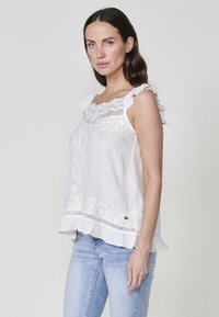 Blouse blanche sans manches en tissu texturé, avec des accents en dentelle, des broderies florales et un ourlet froncé. Design doux et léger.