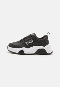 Zapatillas negras con un acabado texturizado, que presentan detalles en blanco y un logo. Diseño de suela gruesa con una entresuela suave y un cordón prominente.