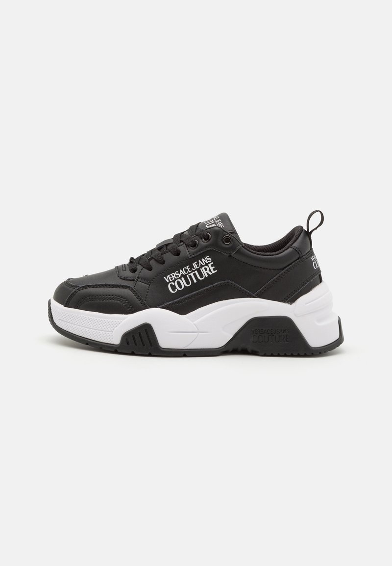 Zapatillas negras con un acabado texturizado, que presentan detalles en blanco y un logo. Diseño de suela gruesa con una entresuela suave y un cordón prominente.
