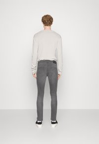 Jeans gris ajustados con detalles de costura, llevados con una camisa de manga larga gris claro y zapatillas negras. La vista trasera muestra los bolsillos y desgastes.