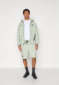 Vaaleanvihreä Nike-urheiluvaatesetti, joka sisältää vetoketjullisen hupullisen hupparin taskuilla, vastaavat shortsit ja valkoisen T-paidan. Sisältää joukkuelogo ja yksityiskohdat.