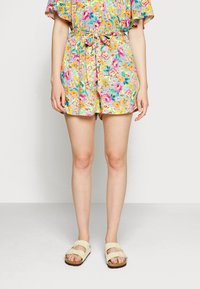Os calções com padrão floral apresentam um fundo azul claro com flores rosa, amarelas e verdes, ajustados com uma cintura atada. Combinados com sandálias bege.