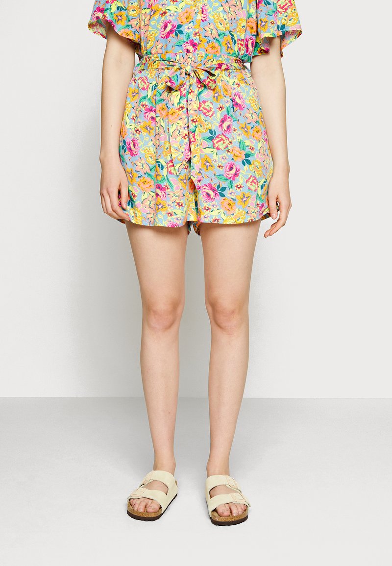 Os calções com padrão floral apresentam um fundo azul claro com flores rosa, amarelas e verdes, ajustados com uma cintura atada. Combinados com sandálias bege.