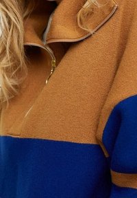 Pull en polaire coloré en blocs marron et bleu avec un col zippé. Texture douce, coupe décontractée et design en contraste sur la poitrine.