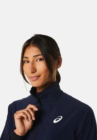 Marineblaue Zip-Jacke aus leichtem Material mit hohem Kragen. Mit einem weißen Logo auf der Brust und einer glatten, strukturierten Oberfläche.